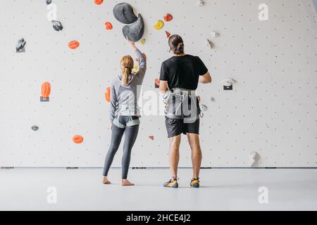 Ganzkörper-Rückansicht ein paar Alpinisten mit Sicherheitsgeschirr, die beim Training im Fitnessstudio neben einer Kletterwand mit bunten Griffen stehen Stockfoto