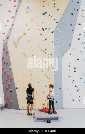 Ganzkörper-Rückansicht ein paar Alpinisten mit Sicherheitsgeschirr, die beim Training im Fitnessstudio neben einer Kletterwand mit bunten Griffen stehen Stockfoto