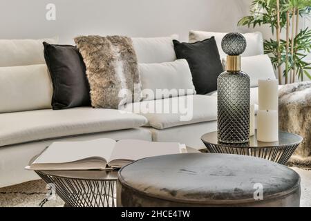 Komfortable weiße Couch mit Kissen auf Teppich neben runden Couchtischen, die im modernen Wohnzimmer mit Kerzen dekoriert sind Stockfoto