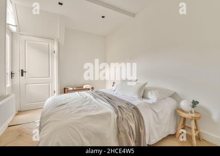 Komfortables Bett mit Kissen und Decke, die an der Wand zwischen grünen Blumen in einem hellen, stilvollen Schlafzimmer mit einem großen Heizkörper im Apartment platziert sind Stockfoto