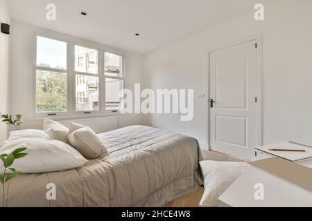 Komfortables Bett mit Kissen und Decke an der Wand zwischen grünen Blumen in hellen stilvollen Schlafzimmer mit Tisch im Apartment platziert Stockfoto