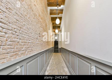 Perspektivischer Blick auf einen leeren schmalen langen Durchgang mit einer weißen Wand und einer Ziegelwand und einer Lampe, die von der Decke zum Treppenhaus führt Stockfoto