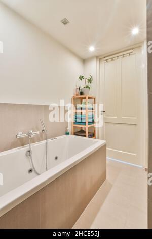 Weiße Badewanne neben einem Regal mit Handtüchern im modernen beleuchteten Badezimmer und im Hintergrund die Zugangstür mit einem Handtuchhalter Stockfoto