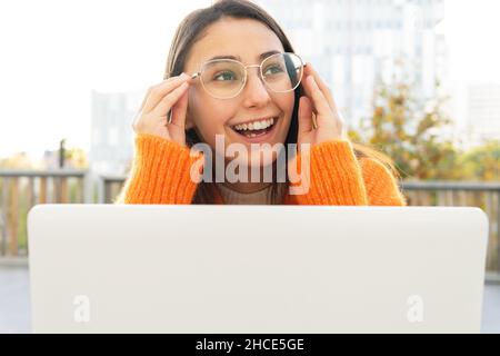 Junge fröhliche Brünette weiblich in hellen Cardigan orange Eingabe auf Laptop-Tastatur während der Arbeit aus der Ferne in der Terrasse Stockfoto