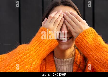 Junge lächelnde Hündin in warmer, orangefarbener Strickjacke aus Wolle, die die Augen bei Tageslicht mit den Händen bedeckt Stockfoto