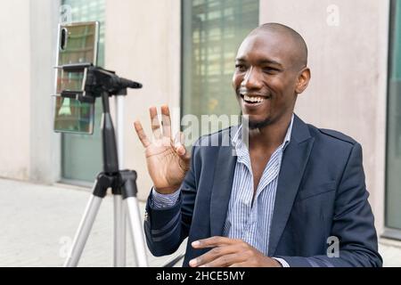 Positiver afroamerikanischer Blogger begrüßt Anzug, während Live-Streaming auf dem Smartphone auf der Straße in der Nähe von Gebäuden in der Stadt Stockfoto