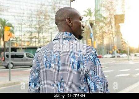 Rückansicht eines kahlen afroamerikanischen Mannes in modischer Kleidung, der auf einer Straße in der Nähe einer Straße in der Stadt wegschaut Stockfoto