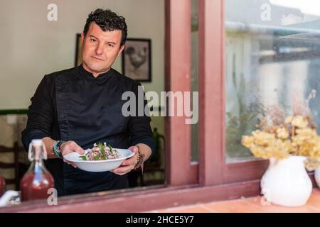 Positiver erwachsener Mann in schwarzem Outfit, der Teller mit leckerem Salat hält, während er im Restaurant steht und auf die Kamera schaut Stockfoto