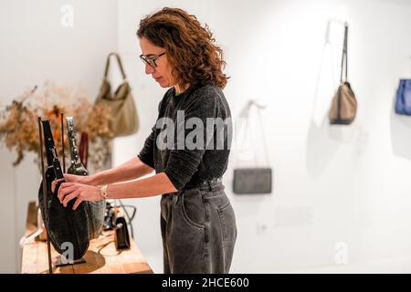 Seitenansicht einer ernsthaften weiblichen Designerin, die neben dem Tisch steht, mit einer Sammlung stilvoller Handtaschen im modernen Lichtstudio Stockfoto