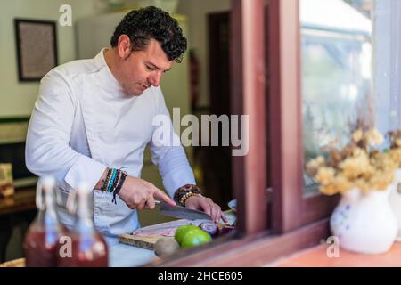 Fokussierter männlicher Koch in Uniform, der Fischfilet an Bord schneidet, während er am Tisch mit verschiedenen Zutaten in der Küche steht Stockfoto