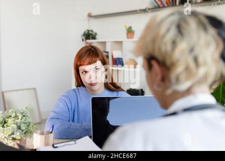 Weibliche Patientin sitzt in der Nähe eines professionellen Arztes mit geöffnetem Ordner, der während der Verabredung in den Laptop zum Surfen geht Stockfoto