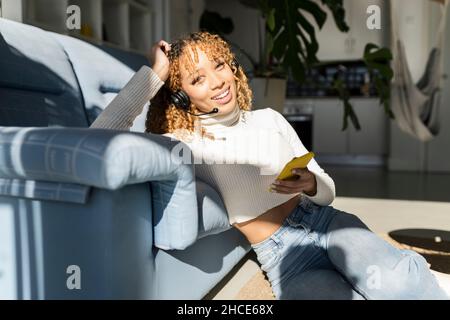 Wunderschöne afroamerikanische Frau mit lockigen Haaren, die ein Headset trägt und ein Smartphone benutzt, während sie auf dem Boden sitzt und die Kamera anschaut Stockfoto