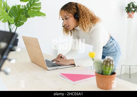 Glückliche afroamerikanische Frau mit lockigen Haaren, die Kopfhörer am Tisch trägt, während sie Videogespräche über einen Laptop führt Stockfoto