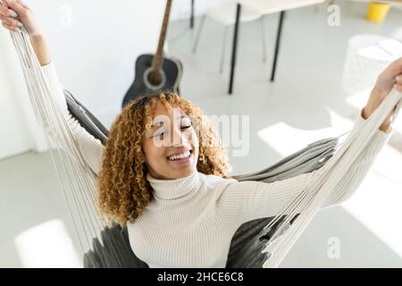 Hoher Winkel der entzückenden schwarzen Dame, die die Augen schließt, während sie in der Hängematte mit erhobenen Armen zu Hause im Sonnenlicht sitzt Stockfoto