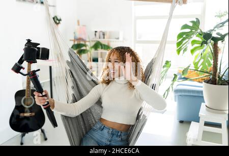 Fröhliche ethnische Musikerin mit Mikrofon begrüßt Anhänger, während sie zu Hause in der Hängematte sitzt und Videos über eine professionelle Kamera dreht Stockfoto