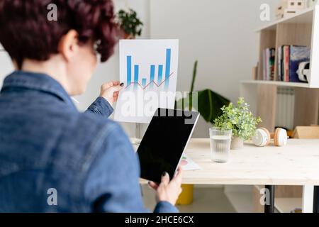 Rückansicht einer anonymen Frau mit einem modernen Tablet mit schwarzem Bildschirm, das am Tisch sitzt und im Home Office Karten auf Papier analysiert Stockfoto