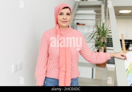 Nachdenkliche Frau mit Krebs im Pullover und rosa Kopftuch, die mit Interesse aus dem Fenster schaut, während sie im hellen Raum steht Stockfoto