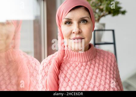 Nachdenkliche Frau mit Krebs im Pullover und rosa Kopftuch, die im hellen Raum auf die Kamera schaut Stockfoto