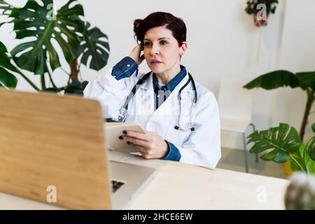 Fokussierte Ärztin in weißer Uniform und Stethoskop, die auf den Bildschirm eines modernen Netbooks schaut, während sie mit dem Buch am Tisch sitzt Stockfoto