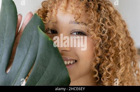 Junge lächelnde afroamerikanische Frau mit lockigen Haaren, die das Gesicht mit grünen Blättern der Monstera-Pflanze bedeckt Stockfoto