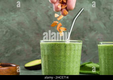 Crop anonyme Person Zugabe von gerösteten Kokosnusssplittern auf grünen Smoothie für Keto-Diät in Glas Stockfoto