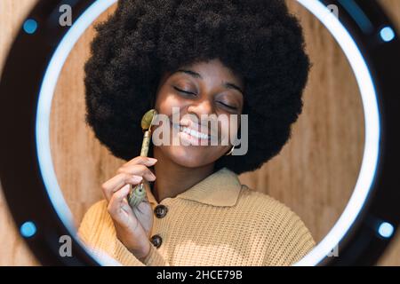 Fröhliche junge afroamerikanische Frau mit Afro-Frisur und geschlossenen Augen massiert Gesicht mit Roller Gesichtsmassager während der Hautpflege Routine zu Hause Stockfoto