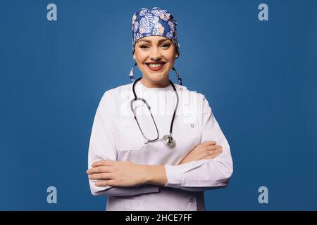 Positive Ärztin in weißer Uniform-Robe und Bandana mit Stethoskop und Blick auf die Kamera mit gekreuzten Armen auf blauem Hintergrund Stockfoto