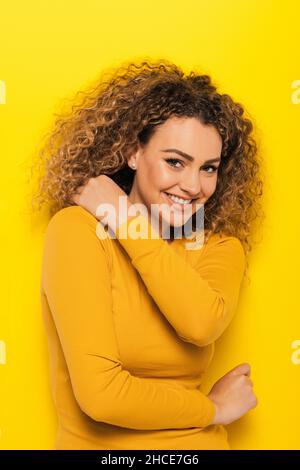 Junge Frau mit lockigen Haaren in hellgelbem Pullover, die auf gelbem Hintergrund die Kamera anschaut Stockfoto