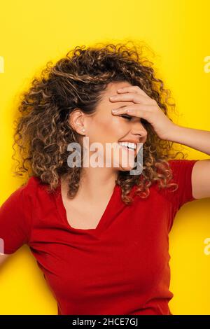 Fröhliche Frau mit lockigen Haaren in rotem T-Shirt lächelt, während sie ihre Augen über gelbem Hintergrund bedeckt Stockfoto