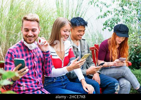 Fröhliche, multiethnische Freunde tragen stilvolle, legere Kleidung, sitzen auf der Bank und surfen auf der Bank im Park durch Mobiltelefone Stockfoto