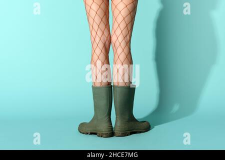 Crop unkenntlich weibliche Modell trägt Netzstrümpfen und schmutzige Gummistiefel im blauen Studio stehen Stockfoto