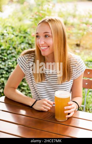 Positive junge blonde Frau in zwanglosem Outfit sitzt am Holztisch mit Glas voller kaltem, schäumenden Lagerbier und lächelt, während sie den Sommer genießt Stockfoto