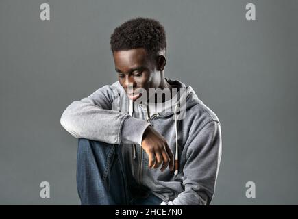 Positives junges schwarzes Männermodell mit dunklem Afro-Haar in trendigen Hoodie und Jeans lächelnd und mit Blick nach unten, während es in studi vor grauem Hintergrund sitzt Stockfoto