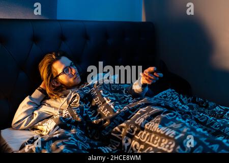 Ein junger Mann, der im Bett liegt und Fernsehen schaut, bevor er einschläft. Lifestyle, nach dunklem Konzept Stockfoto