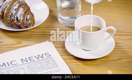 Milch, die auf eine Tasse Kaffee in der Nähe von Zeitungen und Croissants geschüttet wird, Stockbild Stockfoto