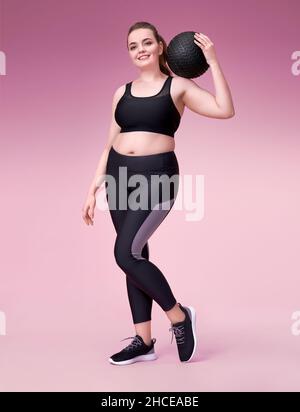 Sportliche Frau mit Medizinball auf der Schulter. Foto des Models mit kurviger Figur in modischer Sportbekleidung auf rosa Hintergrund. Sportliche Motivation und Stockfoto