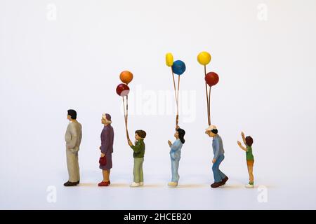 Reihe von Miniaturmenschen unterschiedlichen Alters, einige von ihnen halten bunte Ballons - Generationen Stockfoto
