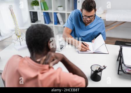 Junge Rehabilitikerin schreibt in einem Notizbuch in der Nähe verschwommener afroamerikanischer Mann, der an Nackenschmerzen leidet Stockfoto