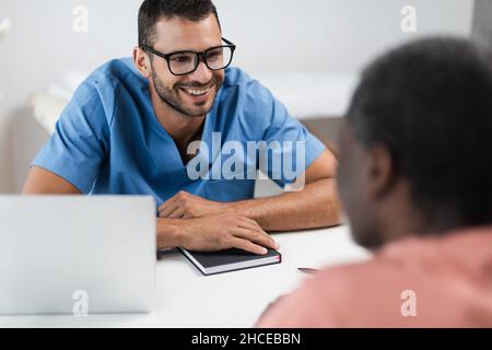 Junger Arzt in Brillen lächelt in der Nähe verschwommener afroamerikanischer Patient im Sprechzimmer Stockfoto