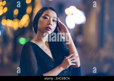 Schönheitsporträt einer jungen asiatischen Frau, die nachts auf der Straße ihr Haar berührt, mit Lichtern im Hintergrund Stockfoto