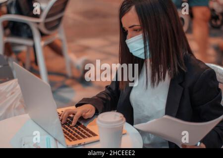 Die Lehrerin trug eine Maske, die auf ihrem Laptop tippte, während sie Untersuchungen im Café korrigierte und Kaffee am Morgen trank Stockfoto