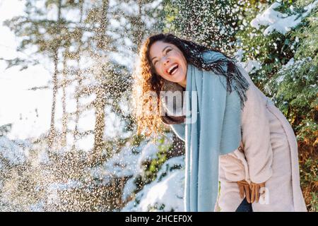 Lächelnd attraktive junge Frau mit langen Haaren haben Spaß unter fallendem Schnee tragen warme Kleidung und blauen Schal in verschneiten Winterwald. Genießen Sie die Natur, Stockfoto