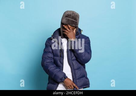 Ein junger trauriger Mann, Student mit warmer Jacke und Pelzhut, der isoliert auf marineblauem Hintergrund posiert. Stockfoto