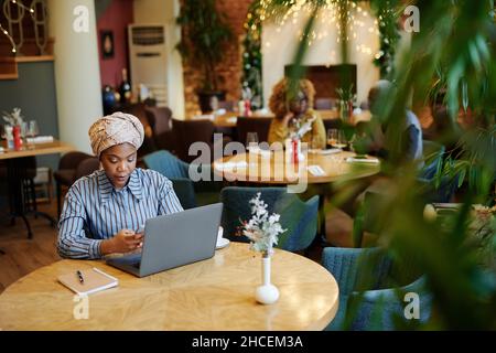 Afrikanische junge Geschäftsfrau, die im Café am Tisch sitzt und einen Laptop für Online-Arbeiten nutzt Stockfoto
