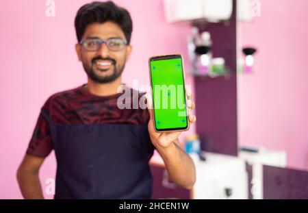 Rack Fokus vom Labrer zum Smatphone, junger Barbier in Arbeitskleidung Showig Handy mit grünem Bildschirm im Salon - Konzept der Werbung, klein Stockfoto