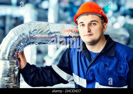 Portrait des jungen Ingenieurs im Helm in der Arbeitswelt in der Fabrik. Gasarbeiter blickt selbstbewusst direkt in die Kamera. Gasausrüstungsmonteur. Nahaufnahme des Mitarbeiters in Schutzkleidung im Heizraum. Heller Hintergrund. Stockfoto