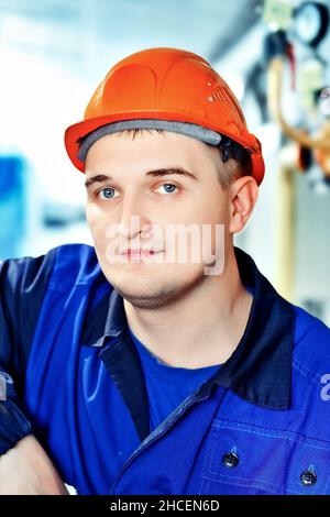 Portrait des jungen Ingenieurs im Helm in der Arbeitswelt in der Fabrik. Gasarbeiter blickt selbstbewusst direkt in die Kamera. Gasausrüstungsmonteur. Nahaufnahme des Mitarbeiters in Schutzkleidung im Heizraum. Heller Hintergrund. Stockfoto