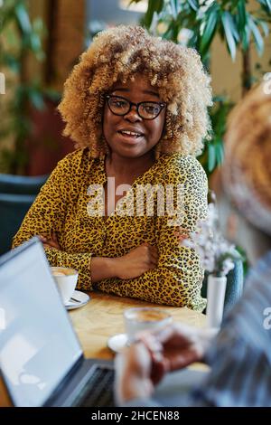 Afrikanische Geschäftsfrau mit lockigen Haaren im Gespräch mit ihrem Kollegen während der Kaffeepause am Tisch im Café Stockfoto