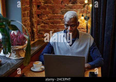 Afrikanischer junger Mann, der online am Laptop arbeitet und Kaffee trinkt, während er im Café am Tisch sitzt Stockfoto