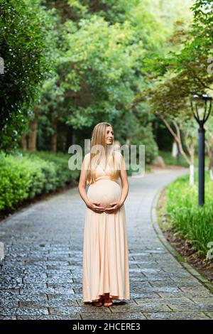 Schwangere Frau in Abendkleid hält Hände auf Bauch im Freien in Bambuswald. Schwangerschaft, Mutterschaft, Vorbereitung und Erwartung Konzept. Wunderschön Stockfoto
